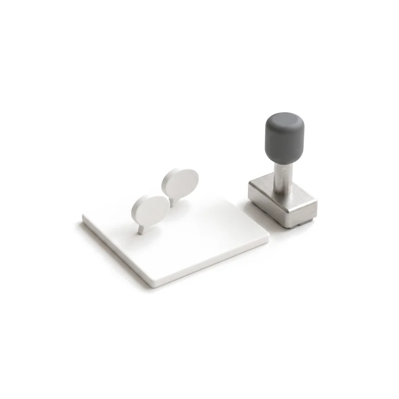 A white ceramic image tile with two ceramic comment pins on it, a steel stamp with a graphite handle beside it.