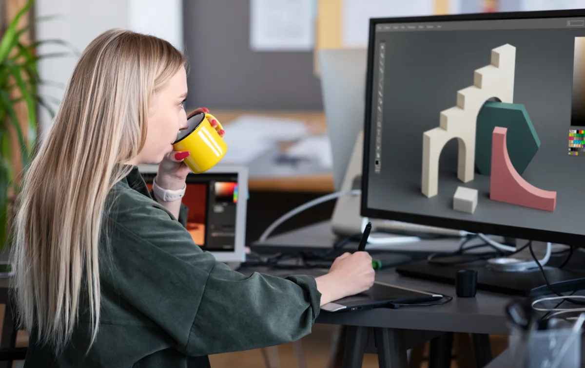 A person holding a yellow mug uses a pen tablet beside a monitor showing a 3D composition.
